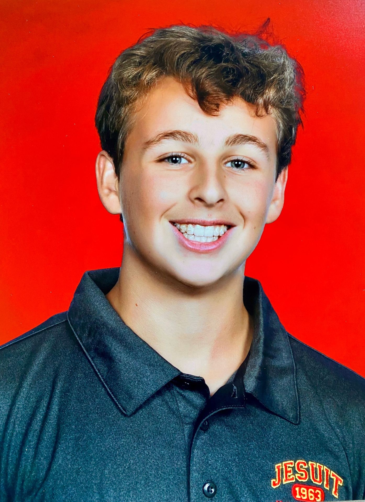 Person wearing a Jesuit 1963 branded black polo shirt against a bright red background.
